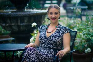 A woman sitting outdoors on a metal chair at a round metal table with intricate perforations. She has light skin, curly hair pulled back, and is smiling warmly. She wears a patterned dress with black and white abstract shapes and a colorful beaded necklace. She also has large spiral-shaped black earrings and a ring on her right hand. The background features greenery, white flowers, and a large stone fountain, suggesting a garden or park setting.